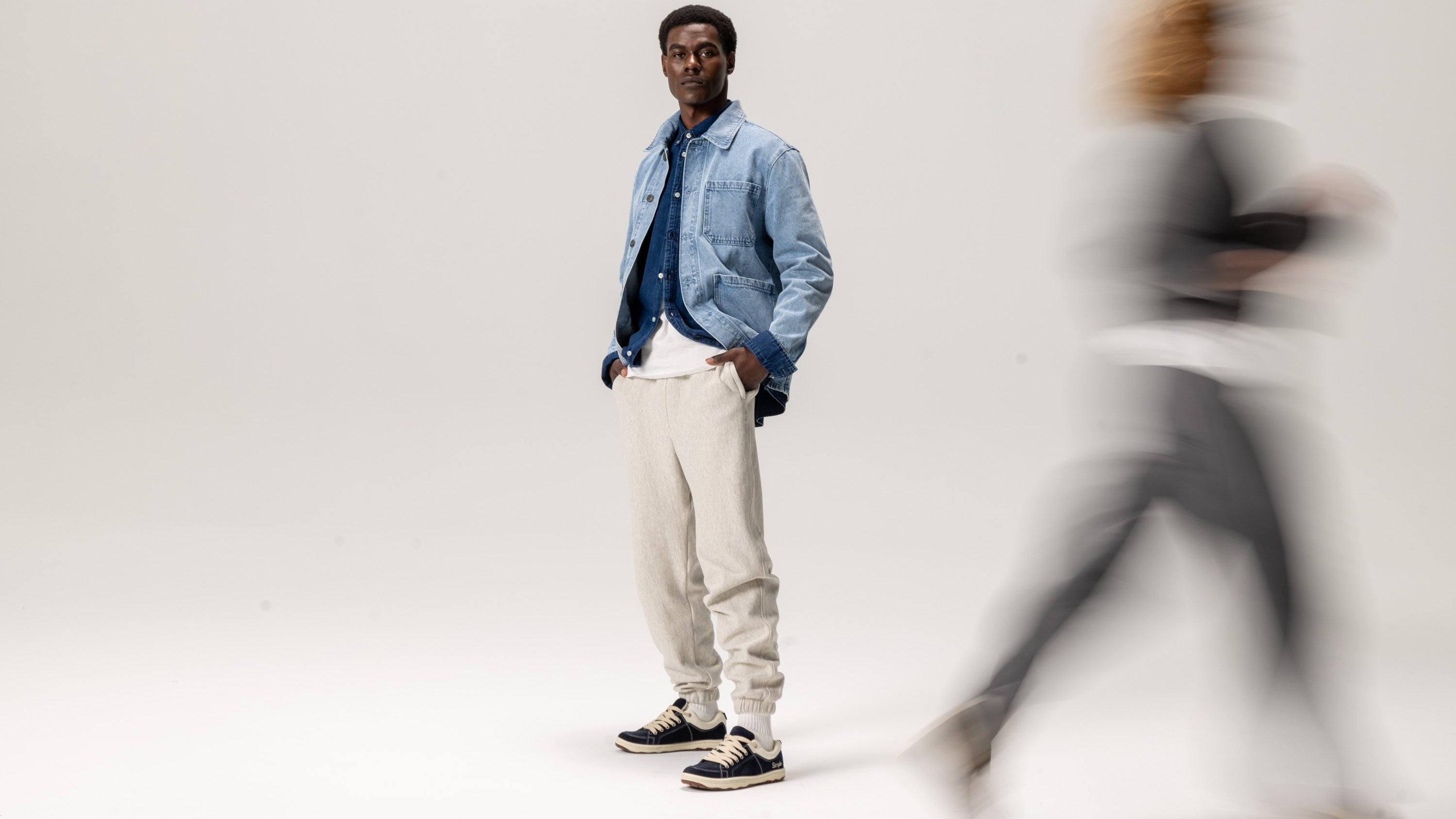 Man in a denim jacket and light pants standing against a white background with a blurred person in the foreground.
