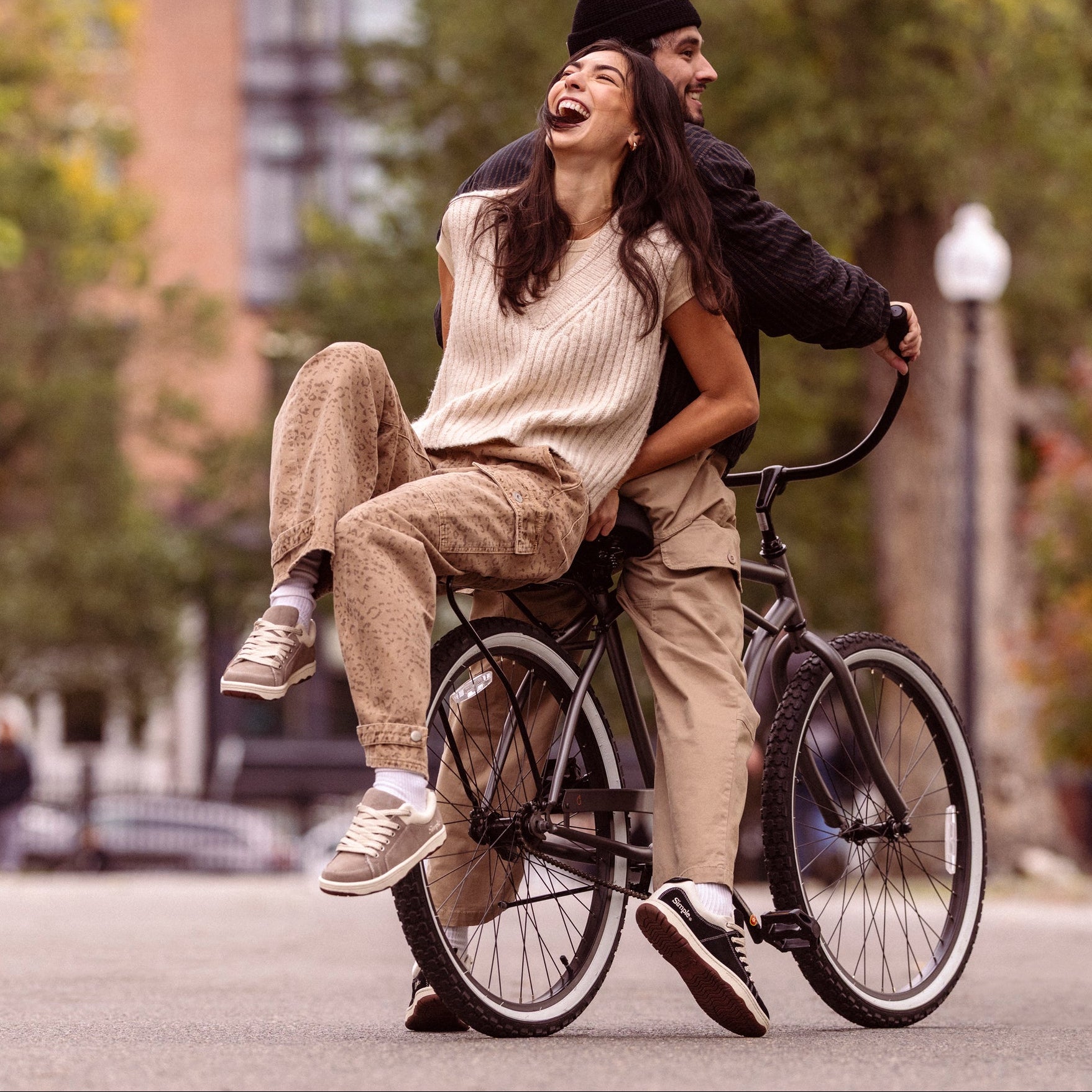 Two people riding a bicycle together on a street with trees and buildings in the background.