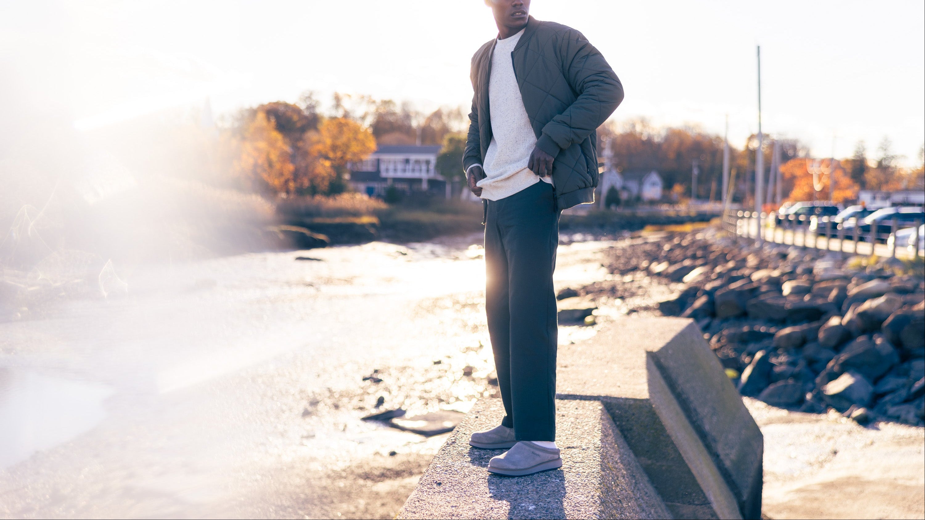 Man standing on a sidewalk by a waterfront with cars and trees in the background