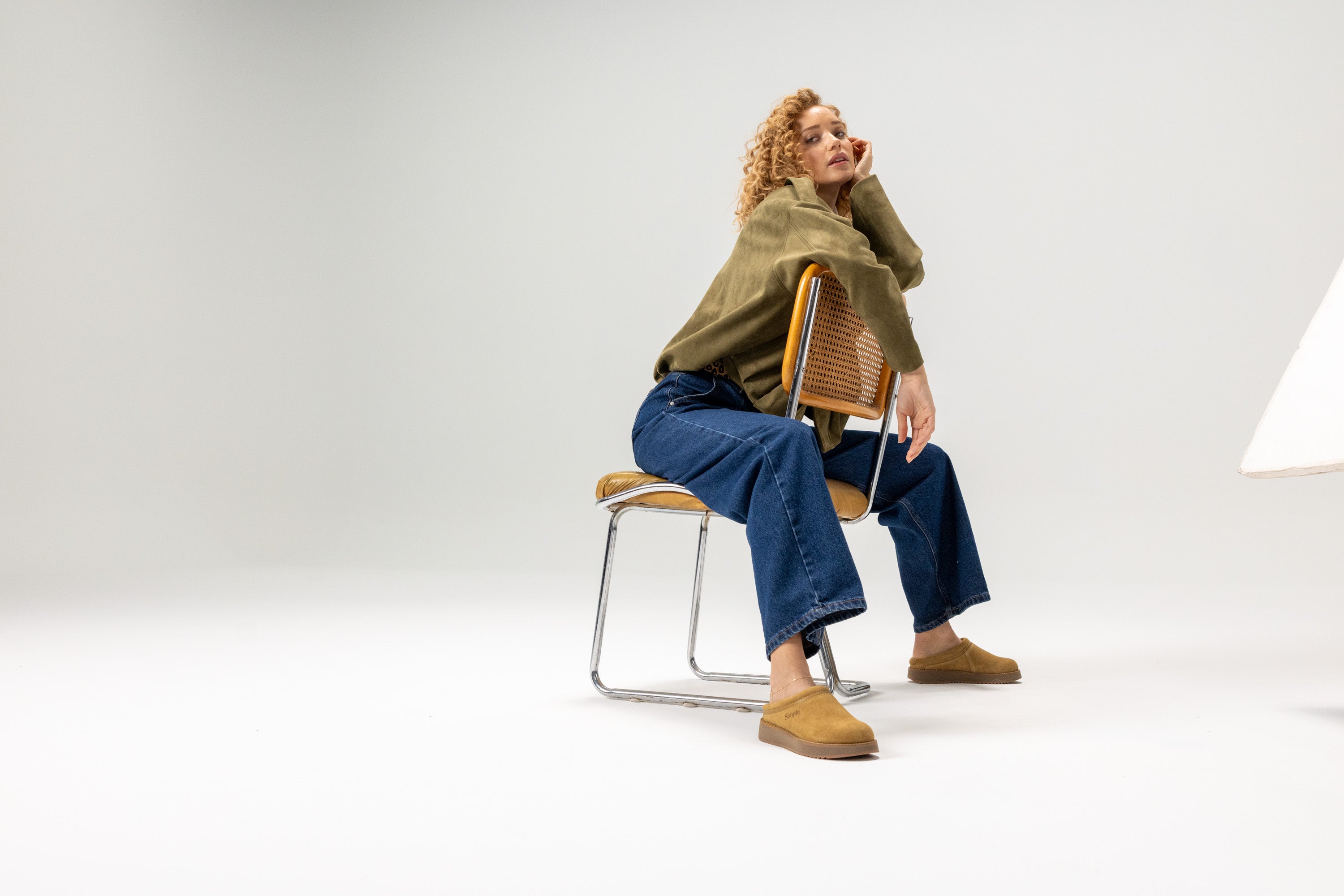 Woman sitting on a chair wearing a green sweater and blue jeans against a white background