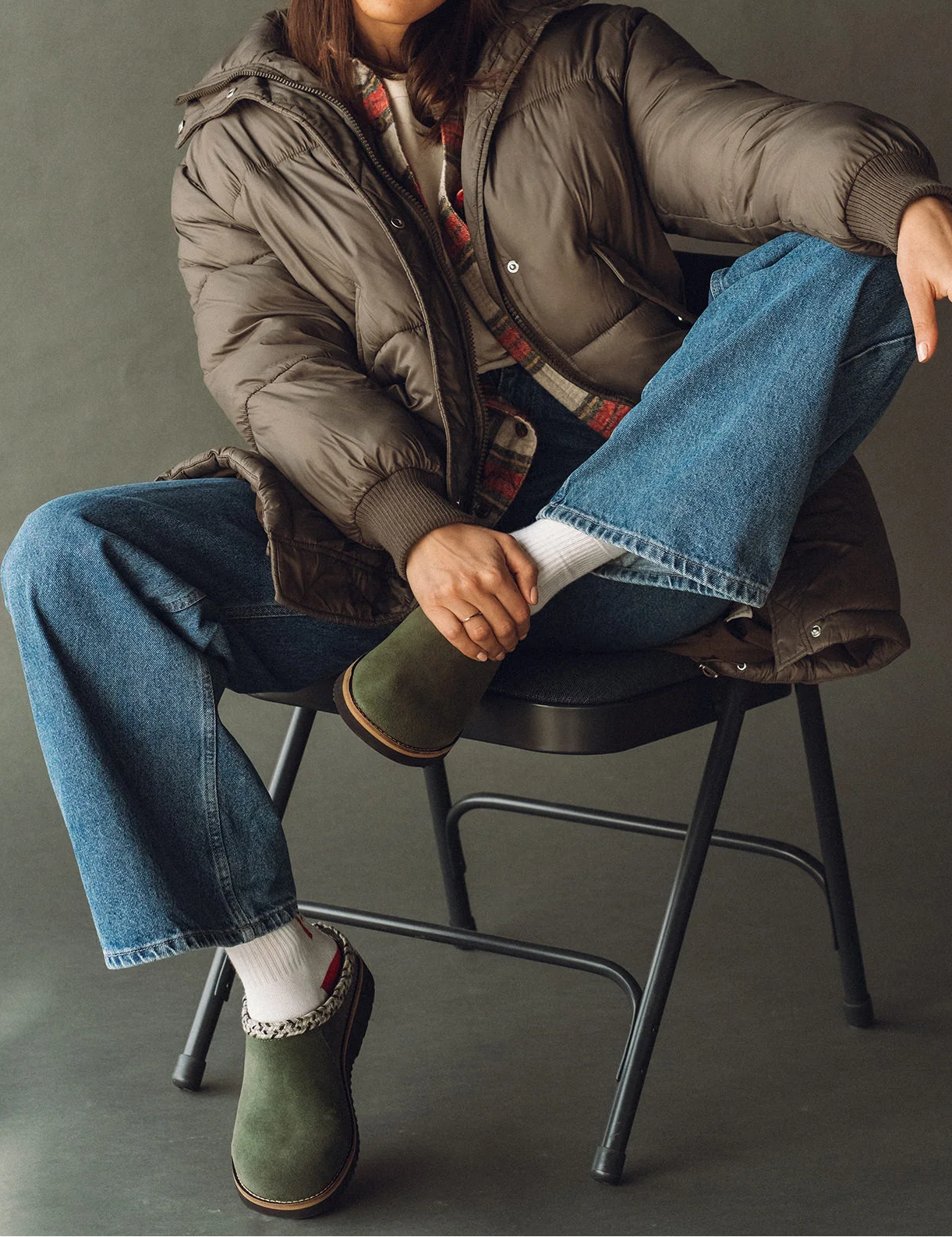 Person wearing a green jacket, blue jeans, and a beanie sitting on a chair against a gray background