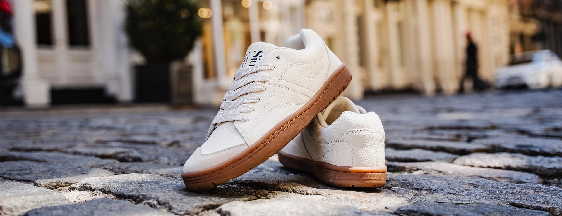 A pair of white low-top sneakers with gum brown soles, photographed outdoors on a cobblestone street with a blurred urban background.