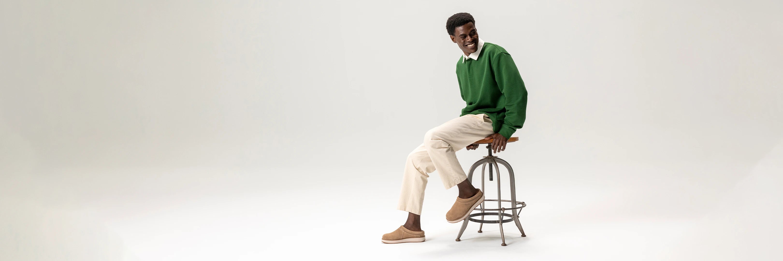 Man sitting on a stool wearing a green sweater and beige pants against a white background