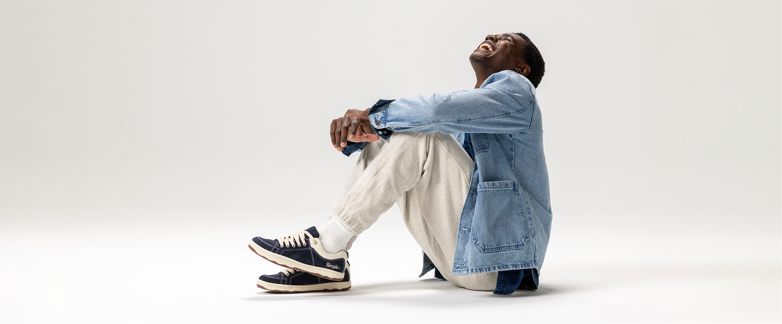 Person sitting on a white background wearing a denim jacket and light pants.