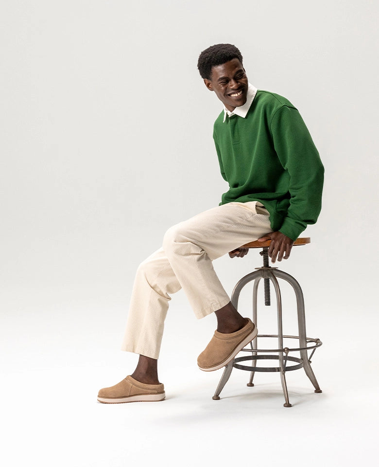 Man sitting on a stool wearing a green sweater and beige pants against a white background