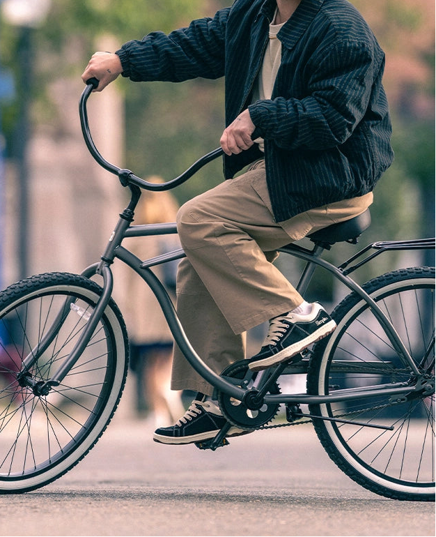 A man riding a black cruiser bicycle outdoors, wearing a black beanie, black pinstriped jacket, beige pants, and black sneakers with white soles.